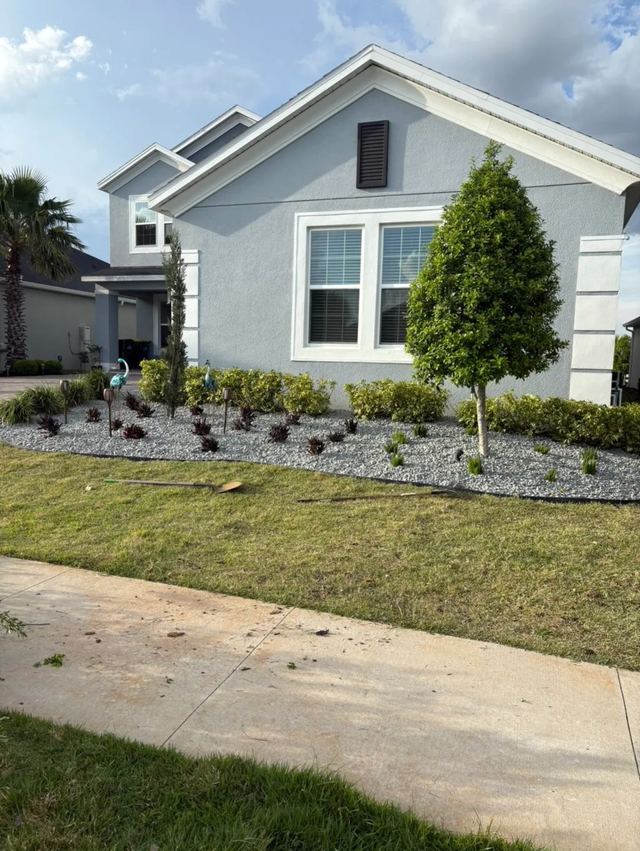 New front-yard bed with white rock, shrubs and a young tree in front of a gray house